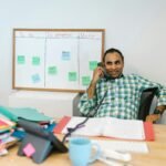Home An adult male in a modern office setting, working at a desk with a task board in the background.
